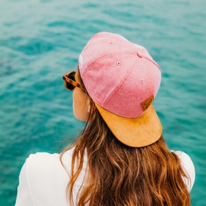 Könnte beinhalten: Eine Frau mit langen braunen Haaren trägt eine rosa und braune Baseballkappe mit braunem Schirm. Sie blickt auf das Wasser.