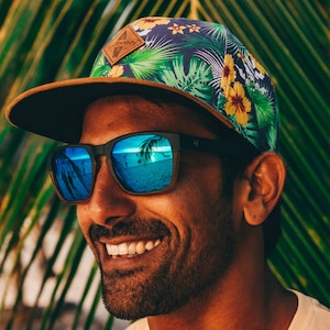 Può includere: Una persona indossa un cappello da baseball con motivo floreale blu e verde, visiera marrone e occhiali da sole blu a specchio. Il cappello ha una toppa in pelle marrone con il testo "Soul Buddy" su di essa.