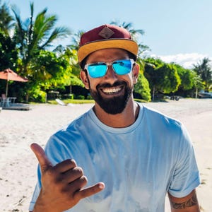 Ein lachender Mann mit Sonnenbrille trägt die Snapback Cap und zeigt mit dem Finger auf die Kamera. Die Cap sitzt lässig, im Hintergrund ist ein Strand zu sehen.