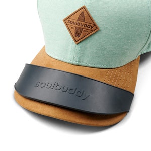 Op de afbeelding: Een mintgroene en bruine baseballpet met een zwart vizier en een bruin leren patch met de tekst "soulbuddy est. 2019" op de voorkant. Het vizier heeft de tekst "soulbuddy" erop.
