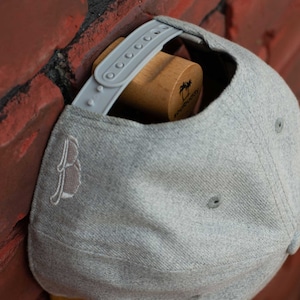 Puede incluir: Gorra de béisbol gris con un logotipo bordado marrón en la parte delantera y una correa gris con una hebilla de plástico. La gorra se muestra en un soporte para sombreros de madera con el texto "Soundbuddy" impreso en el lateral.