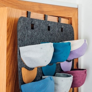 Pode incluir: Um organizador de chapéus de feltro cinza com vários bolsos, pendurado em uma porta de madeira. O organizador contém vários bonés de beisebol em tons de cinza, azul, roxo e bordô. O organizador tem ganchos de metal preto para pendurar.