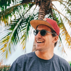 Può includere: Un uomo con un cappello da baseball rosso e marrone con una visiera marrone chiaro, occhiali da sole grigi e una maglietta blu. Sta sorridendo e guardando a destra.