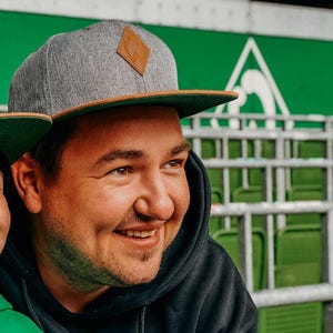Może przedstawiać: Mężczyzna w szarej i brązowej czapce z daszkiem z brązową skórzaną naszywką. Uśmiecha się i patrzy w prawo. Ma na sobie czarną bluzę z kapturem.