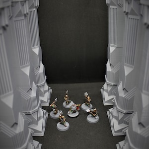 May include: A group of six miniature figures, painted in green, brown, and gold, are arranged in a circle on a grey surface. The figures are standing in front of two tall, grey, rectangular structures.