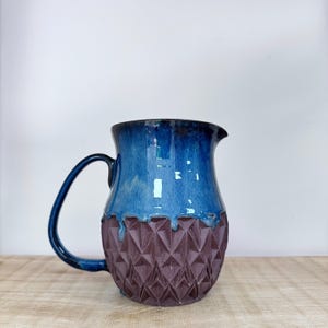 Peut inclure: Pichet en céramique fait à la main avec un dessus émaillé bleu foncé et un motif géométrique texturé de couleur bordeaux foncé en bas. Le pichet a une anse incurvée et un bec verseur.