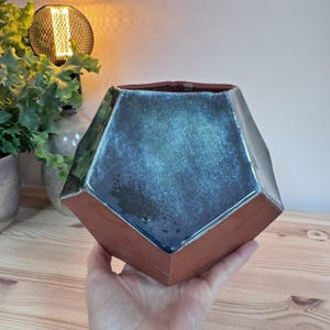 Può includere: Un vaso da fiori in ceramica geometrico con una smaltatura blu scuro e un bordo in terracotta. Il vaso ha un design unico e sfaccettato.