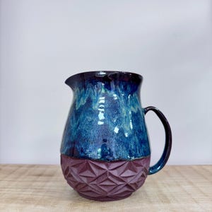 Peut inclure: Pichet en céramique fait à la main, avec un glaçage bleu foncé et violet. La moitié supérieure du pichet est bleu brillant, tandis que la moitié inférieure présente un motif géométrique texturé en violet foncé. Il a une anse incurvée.