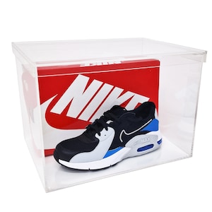 May include: A clear acrylic shoe box containing a red and white Nike shoe box. The shoe box features the Nike logo and the word "Nike" printed on it. A black, white and blue Nike Air Max shoe is inside the box.