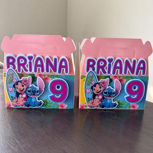 May include: Two pink party favor boxes with the name "Briana" in blue letters. Each box features a colorful illustration of Lilo and Stitch, a surfboard, and the number "9". The boxes are on a dark brown surface.