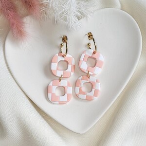 May include: Pink and white checkered square earrings with gold-colored hoops. The earrings are displayed on a white heart-shaped plate, with pink and white decorative elements in the background. The earrings have a geometric design.