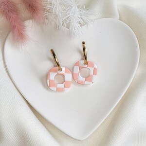 May include: A pair of gold hoop earrings with square, checkerboard-patterned charms in white and light pink. The earrings are displayed on a white, heart-shaped plate, with pink and white decorative elements in the background.