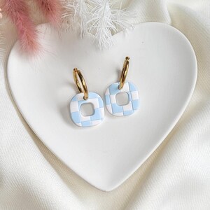 May include: A pair of gold hoop earrings with square, light blue and white checkered pendants. The earrings are displayed on a white heart-shaped dish, with pink and white decorative flowers in the background.