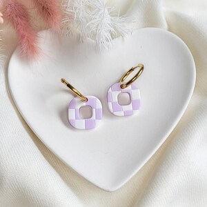 May include: A pair of square-shaped earrings with a checkerboard pattern in white and light purple. The earrings have gold-colored hoop closures. The earrings are displayed on a white heart-shaped plate, with pink and white decorative flowers in the background.