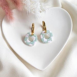 May include: A pair of gold hoop earrings with square, mint green and white checkered pendants. The earrings are displayed on a white heart-shaped dish, with pink and white decorative elements in the background.