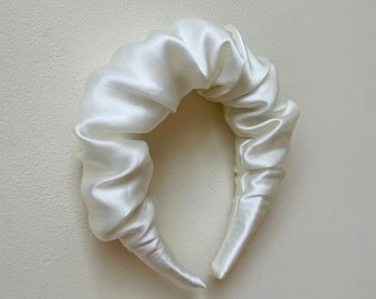 May include: A white satin headband with a gathered, ruffled design. The headband is made of soft, luxurious fabric and is perfect for adding a touch of elegance to any outfit.