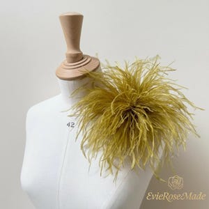 May include: A vibrant mustard yellow ostrich feather accessory attached to a white fabric mannequin torso. The feathers are long, wispy, and fan out in a circular shape. The mannequin features a wooden neck piece and the number 42 printed on it.