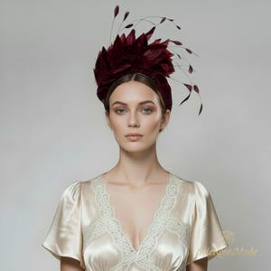 Pode incluir: Um toucado fascinator em bordeaux profundo com pétalas de veludo em camadas e detalhes delicados de penas finas que se estendem para fora. O toucado é usado com uma blusa de cetim creme de manga curta com acabamento em renda no decote.