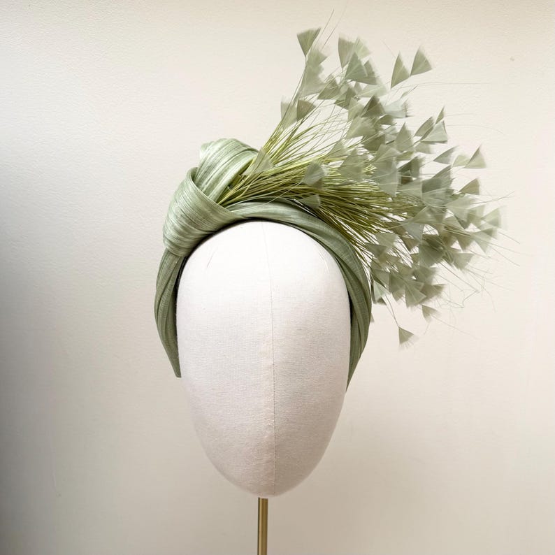 May include: A sage green headband featuring a knotted fabric design on one side. The accessory is adorned with a cluster of delicate, fan-shaped feathers and thin, wispy stalks in a matching light green hue, extending outward from the knot.