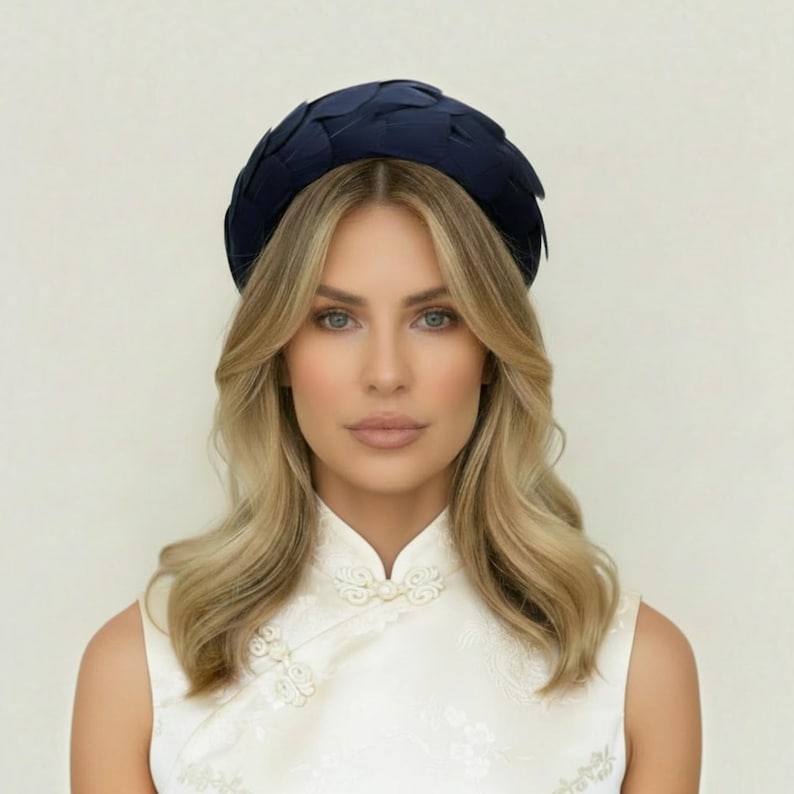 May include: A dark navy blue headband featuring a woven, layered petal design. The headband has a rounded, structured shape that sits atop the head, showcasing a smooth, matte finish with overlapping fabric segments creating a textured appearance.