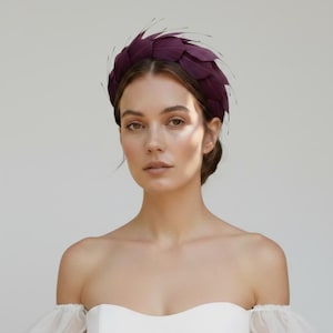 Puede incluir: Una diadema de color ciruela intenso adornada con plumas superpuestas dispuestas en una formación circular similar a una corona. Las plumas son puntiagudas y se extienden ligeramente hacia afuera, creando una apariencia texturizada en forma de abanico.