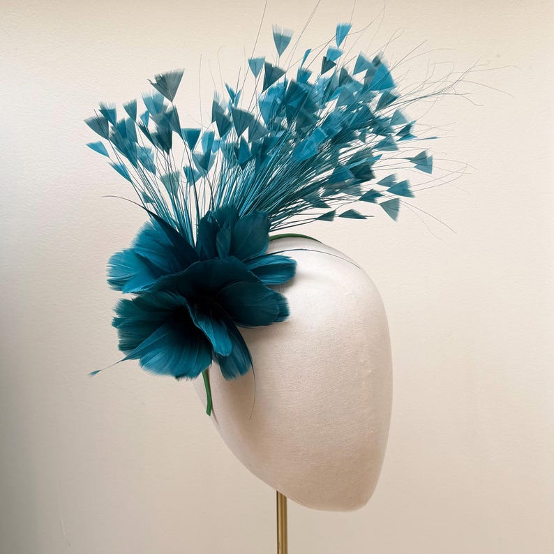May include: A teal fascinator headpiece featuring a dense cluster of soft, layered feathers at the base, accented by a tall, sweeping arrangement of thin, wire-like stems topped with small, fan-shaped teal feather tips extending outward.