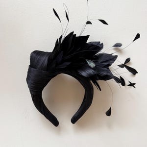 May include: A black headband featuring a wide, wrapped fabric band and an elaborate arrangement of black feathers. The design includes dense, layered feathers on one side, accented by several thin, wispy stems extending outward from the main cluster.