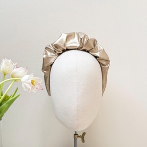 May include: A gold-colored headband made of faux leather with a gathered, ruffled design. The headband is displayed on a white mannequin head.