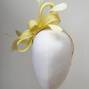 May include: A bright yellow fascinator headpiece featuring a woven straw-like texture with structured loops and delicate, pale yellow feathers extending upwards and outwards, all attached to a thin, matching yellow headband.