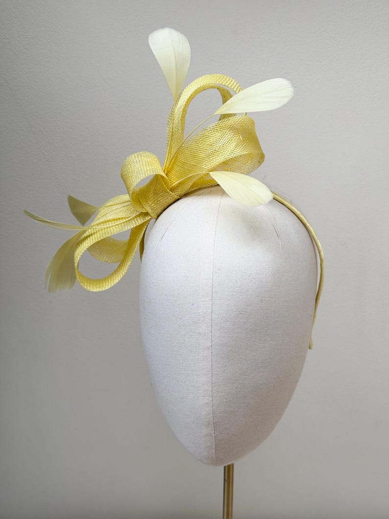 May include: A bright yellow fascinator headpiece featuring a woven straw-like texture with structured loops and delicate, pale yellow feathers extending upwards and outwards, all attached to a thin, matching yellow headband.