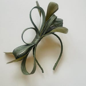May include: Olive green headband with a decorative bow and feather accents. The bow is made of woven material and features looped details. The feathers are a similar shade of green. A stylish hair accessory for special occasions.