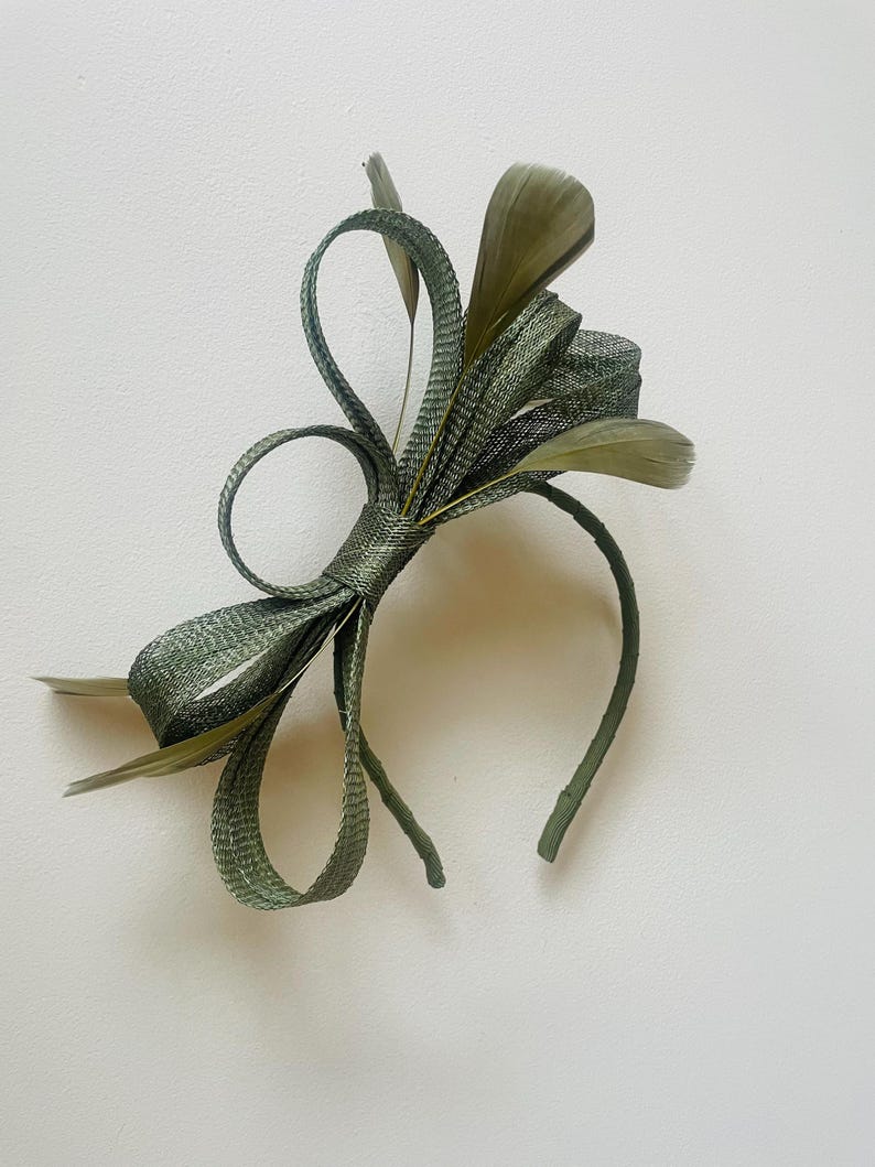 May include: Olive green headband with a decorative bow and feather accents. The bow is made of woven material and features looped details. The feathers are a similar shade of green. A stylish hair accessory for special occasions.