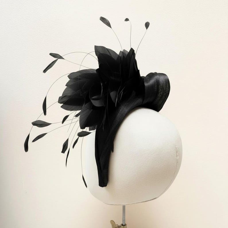 May include: A black fascinator headpiece featuring a structured base and an arrangement of layered black feathers. The design includes thin, delicate stems extending outward, each tipped with a small, singular black feather accent.