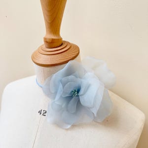 Op de afbeelding: Een lichtblauwe bloemenversiering van stof met een lichtgroen hart, getoond op een wit mannequin.