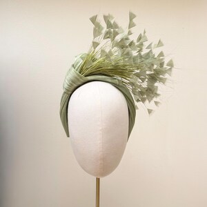 May include: A sage green headband featuring a knotted fabric design on one side. The accessory is adorned with a cluster of thin, radiating stems topped with small, triangular, fan-shaped accents in a matching light green color.