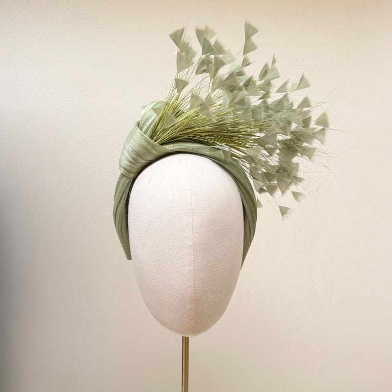 May include: A sage green headband featuring a knotted fabric design on one side. The accessory is adorned with a cluster of thin, radiating stems topped with small, triangular, fan-shaped accents in a matching light green color.