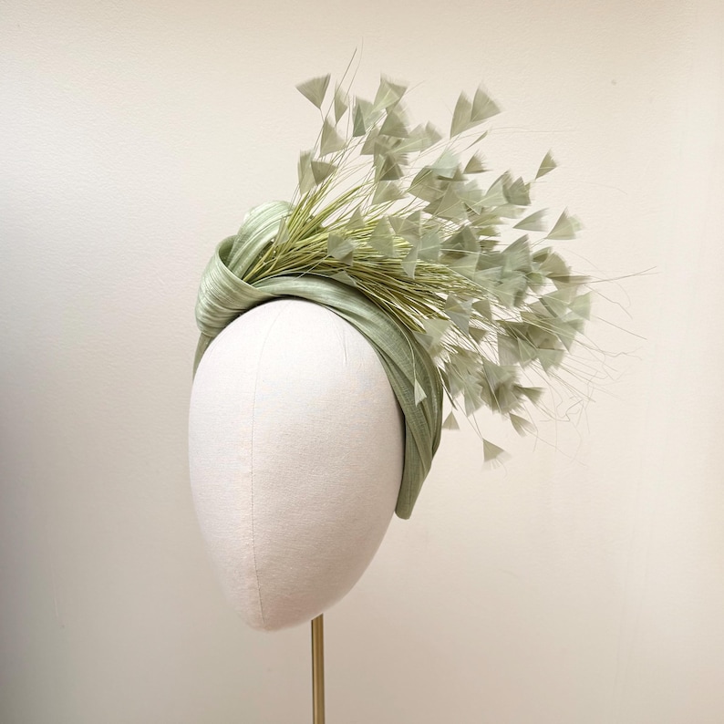 May include: A sage green headband featuring a knotted fabric band and an arrangement of delicate, fan-shaped feather accents. The decorative elements extend upward and outward from the side, creating a textured, layered appearance.