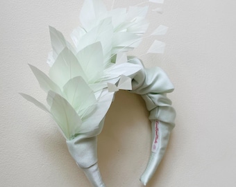 May include: A light green satin headband with a large, white feather embellishment. The headband is tied in a knot at the back and has a small label that says "Darling Miss."