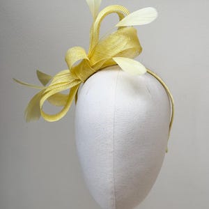 May include: A bright yellow fascinator headpiece featuring a woven straw-like texture with structured loops and delicate, pale yellow feathers extending upwards and outwards, all attached to a thin, matching yellow headband.