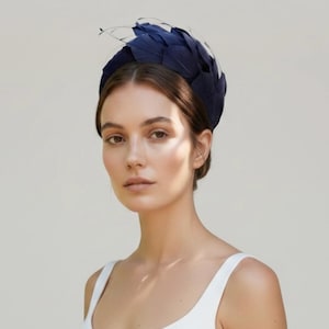 May include: A dark navy blue headpiece featuring a dense arrangement of layered feathers. The accessory is structured in a rounded, crown-like shape that sits atop the head, showcasing a deep, uniform color and a textured, overlapping feather design.