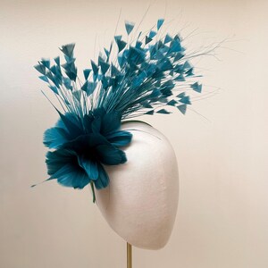 May include: A teal fascinator headpiece featuring a large, layered feather flower at the base with numerous thin, radiating stems topped with small, delicate teal feather tufts extending outward in a fan-like arrangement on a display form.