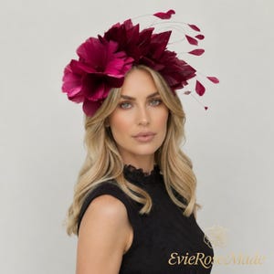 May include: A deep magenta feather fascinator headpiece featuring a dense arrangement of layered feathers and thin, delicate wire stems with small feather accents extending outward, creating a voluminous and textured floral-like appearance.