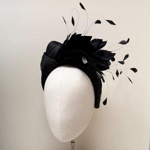 May include: A black headpiece featuring a wide, structured band with a textured, pleated finish. The design is adorned with a cluster of dark feathers and thin, delicate stems extending outward, creating a layered and dimensional appearance.