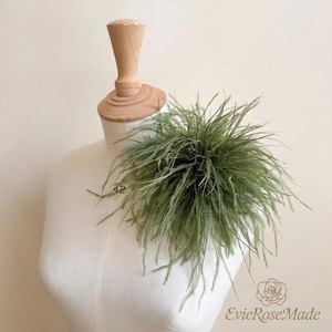 May include: A sage green feather accessory featuring long, wispy, and delicate plumes arranged in a dense, circular cluster. The accessory is displayed on a cream-colored fabric mannequin torso with a wooden neck piece and base.