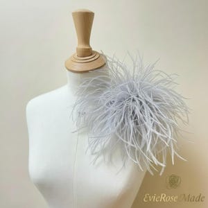 May include: A light gray ostrich feather accessory attached to a white mannequin torso. The feathers are long, thin, and radiate outward from a central point, creating a voluminous, circular shape with a soft, wispy texture and appearance.