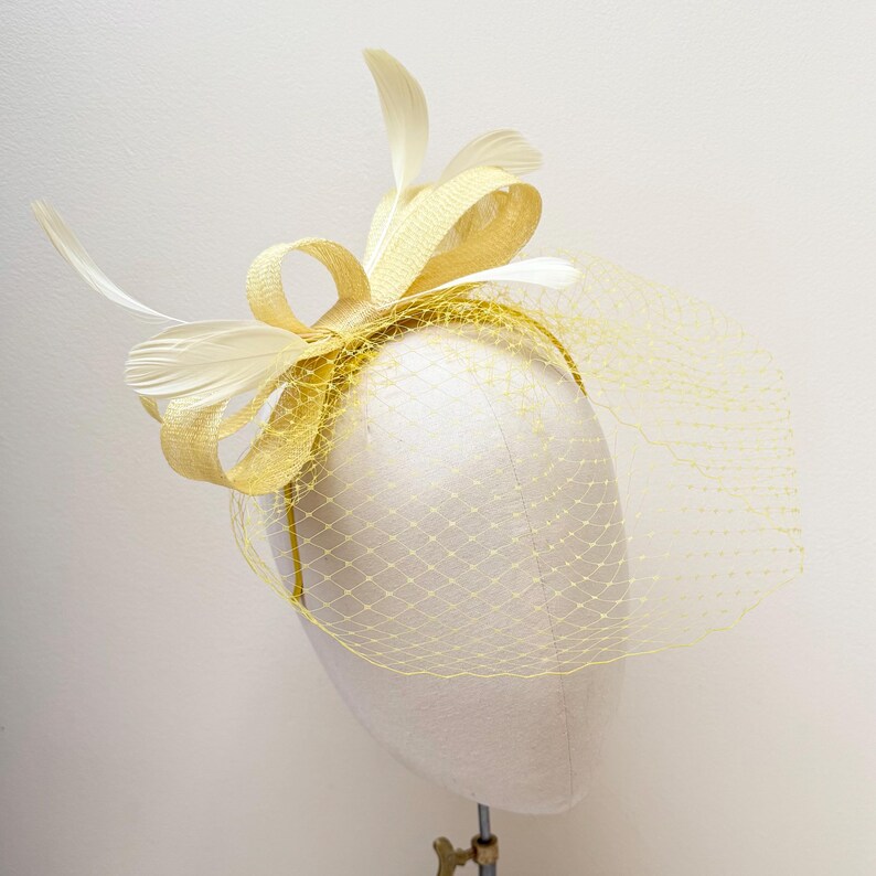 May include: A pale yellow fascinator headpiece featuring a structured bow made of woven material, accented with several light cream-colored feathers. A fine, matching yellow netting veil extends from the base, covering the front of the headpiece.