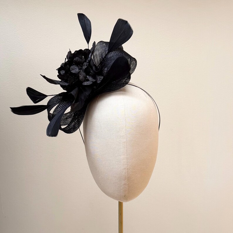 May include: A black fascinator with a floral arrangement and feathers, mounted on a thin headband. The headpiece features a large, dark flower and several black feathers, creating a dramatic and elegant look. The accessory is displayed on a mannequin head.