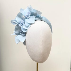 May include: A pale blue headband adorned with fabric flowers. The flowers are clustered on one side of the headband, creating a stylish look. The headband is displayed on a mannequin head.