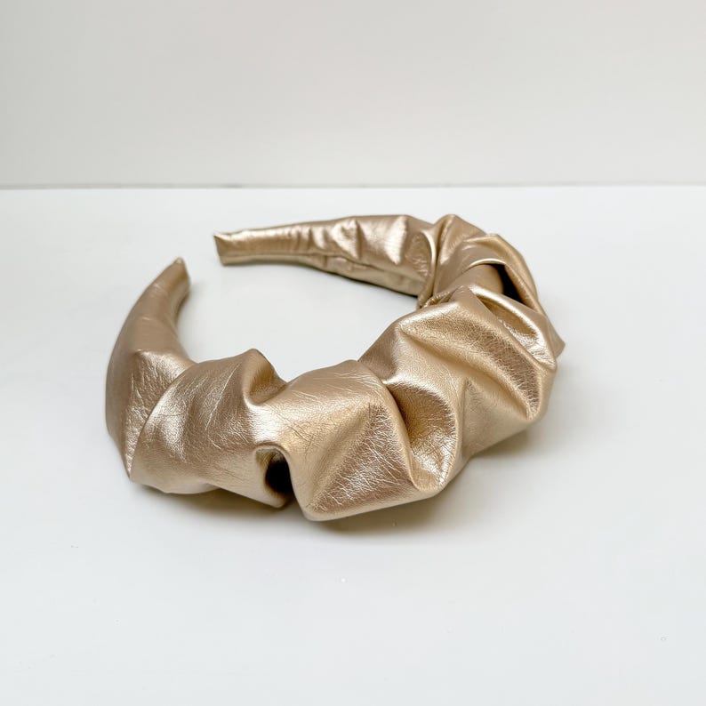 May include: A gold-colored headband made of gathered fabric. The headband is a simple, elegant design.