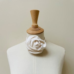 May include: A white satin rose flower, tied into a bow, is displayed on a white mannequin bust.